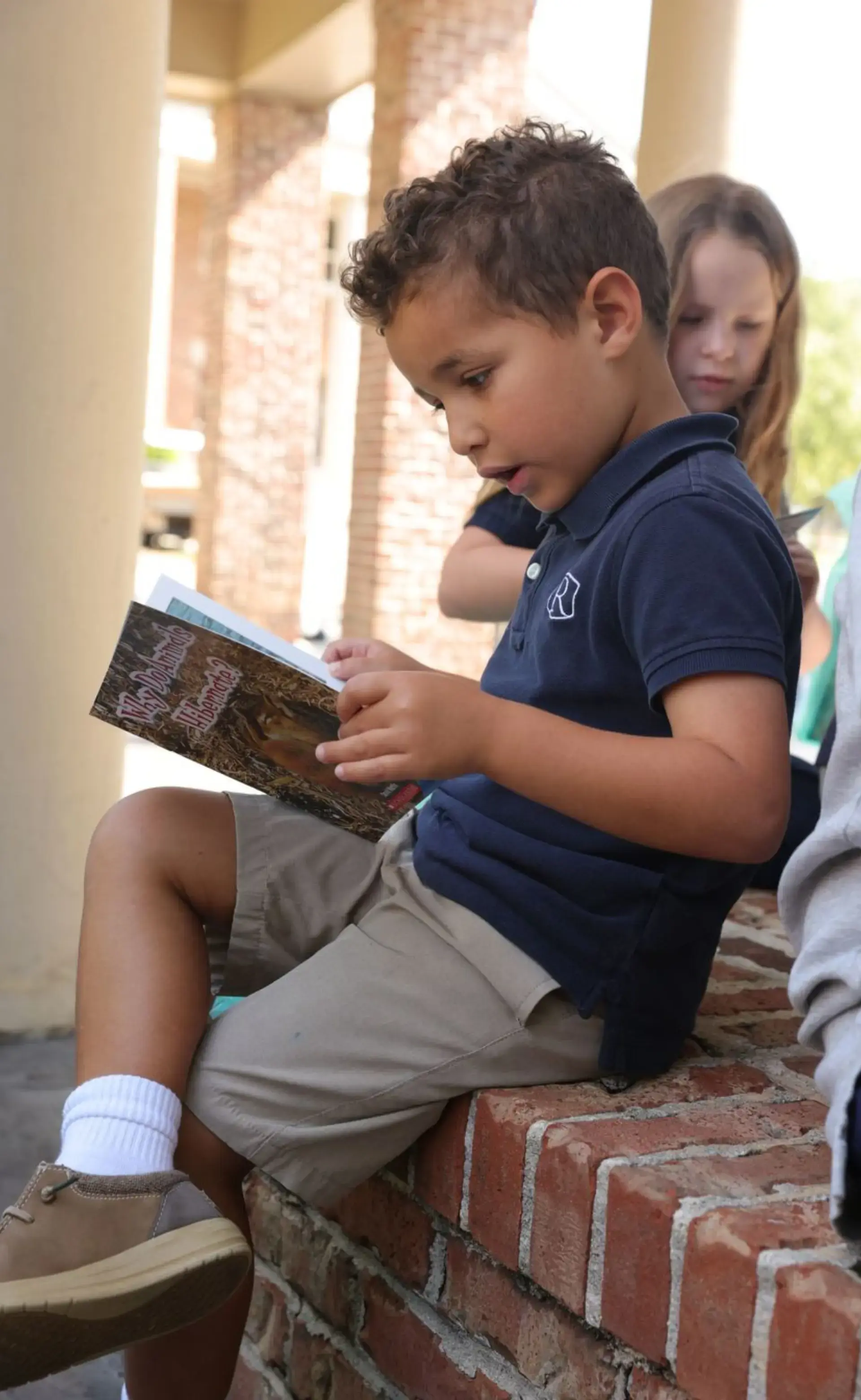 Young student sits outdoors reading a book while classmates gather nearby.