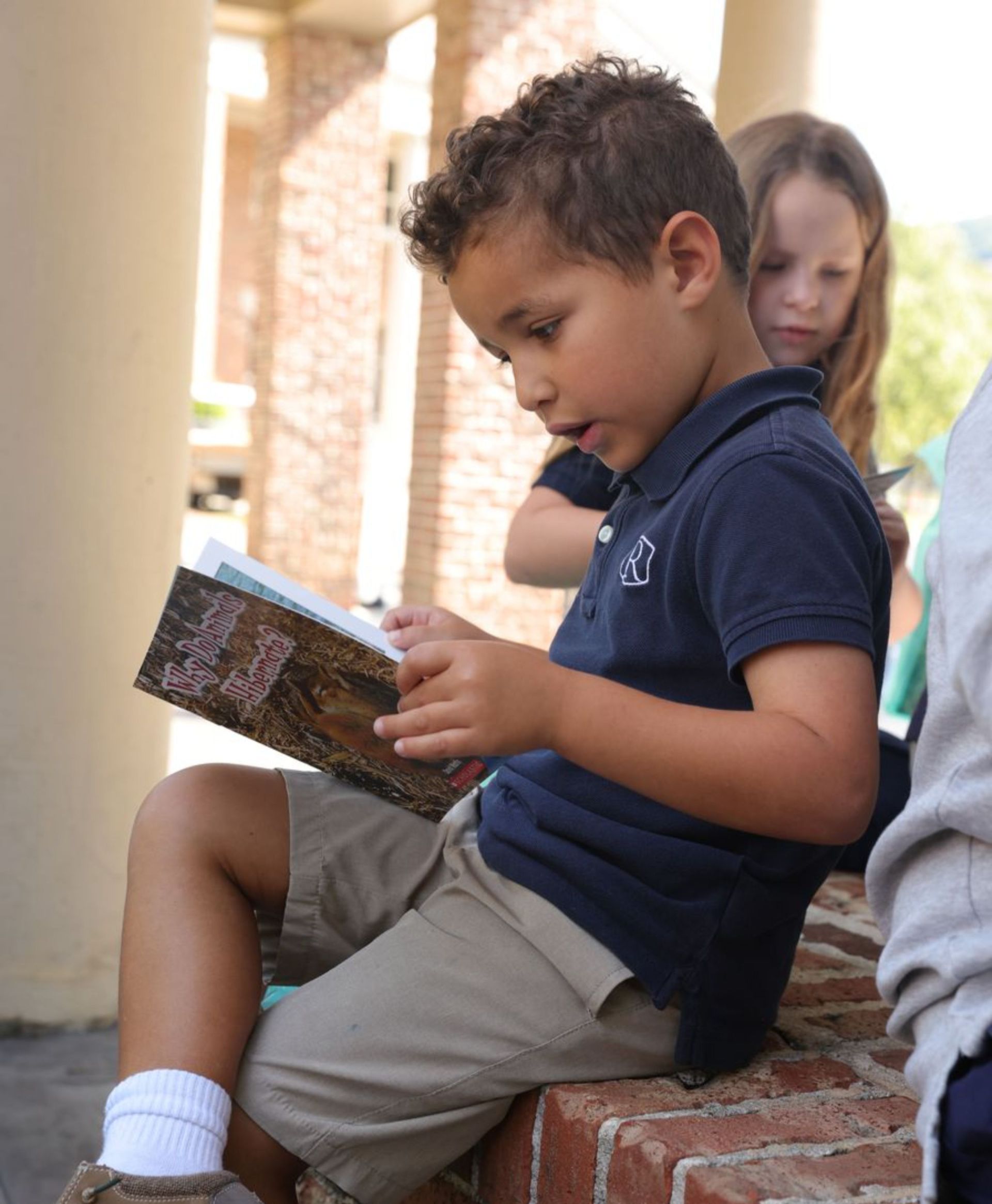 A Randolph Lower School student sits on campus reading a book, focused and engaged in learning.