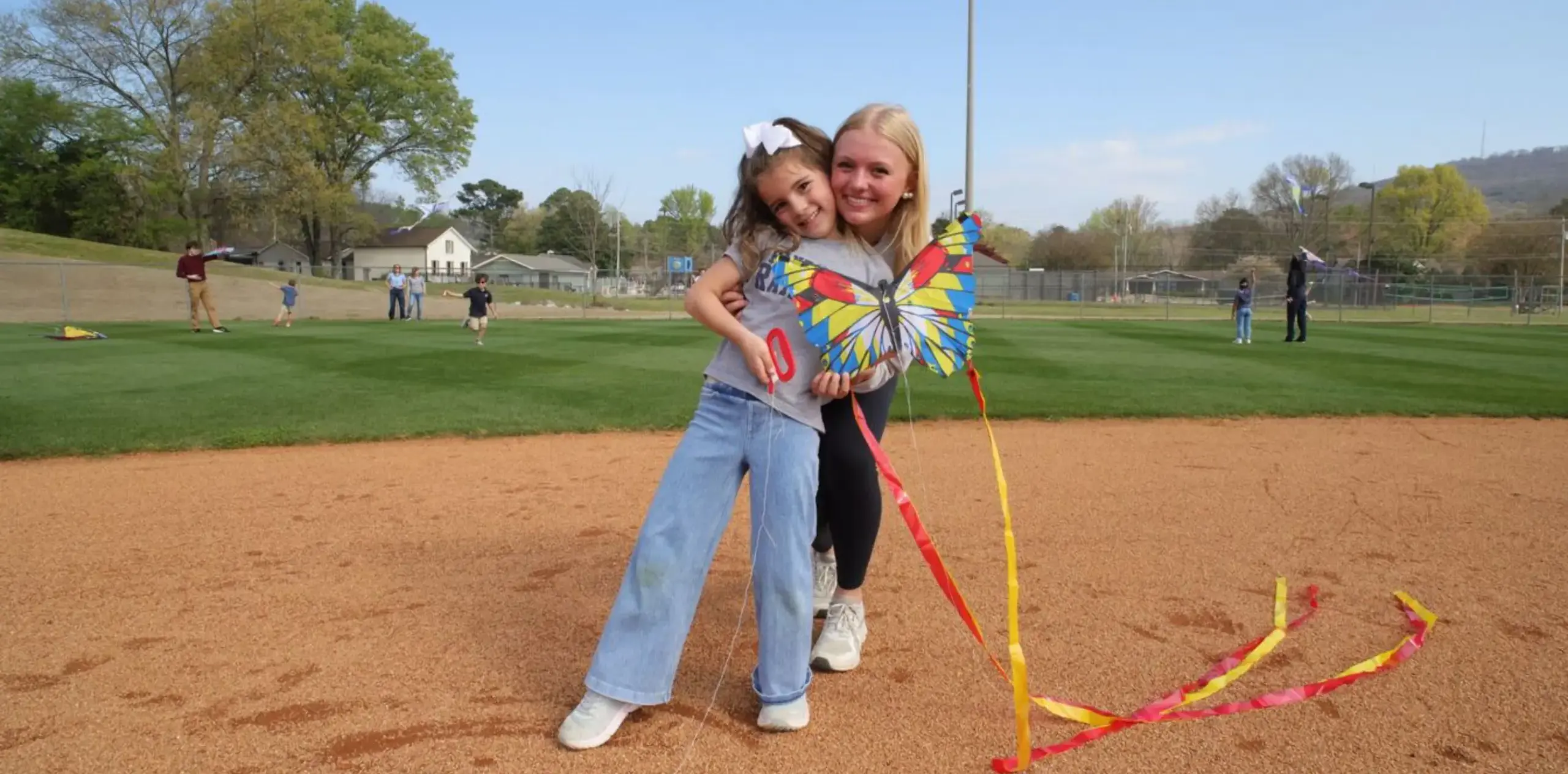 Two smiling students hug while holding a colourful butterfly kite on a sunny field during an outdoor school activity.
