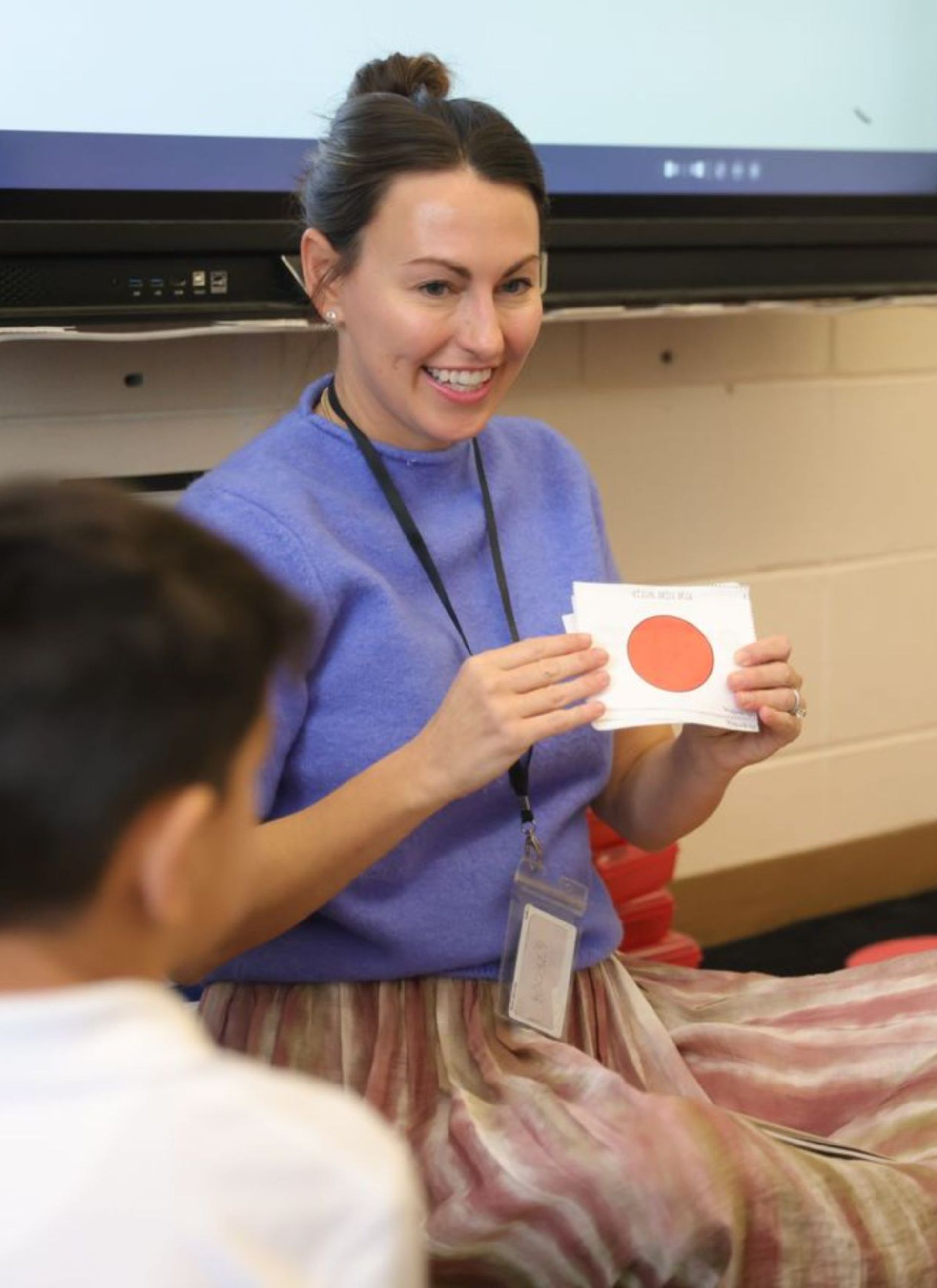 Teacher leading a classroom lesson while holding up a flashcard with a red circle shape.