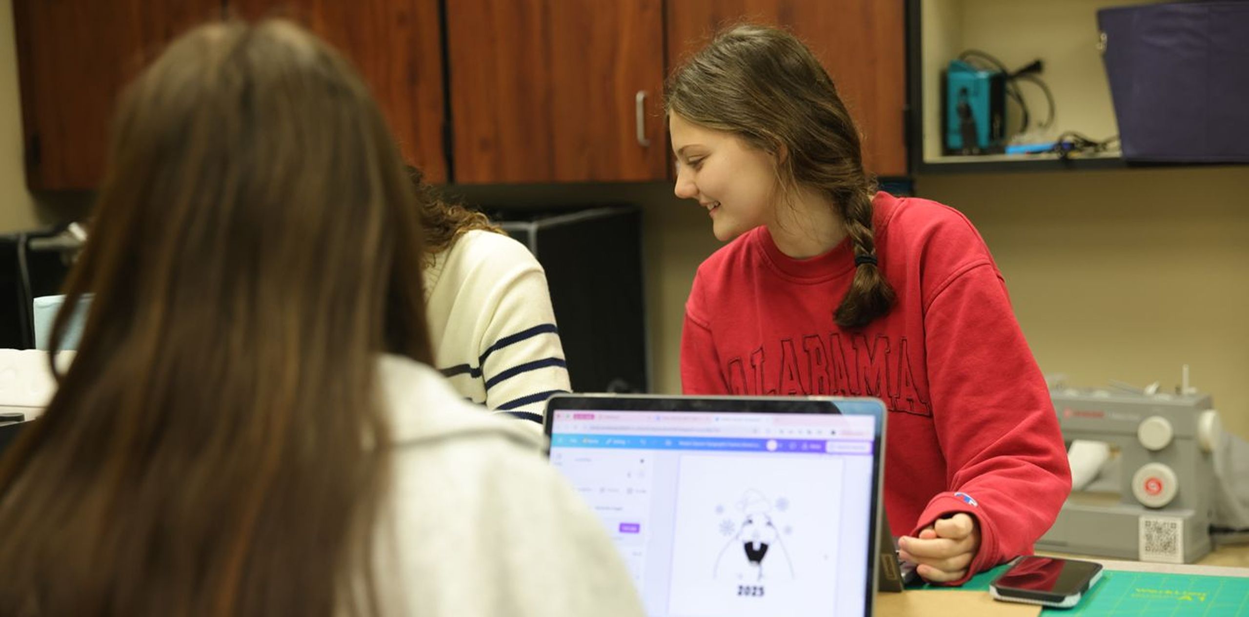 Students collaborate on a creative design project using a laptop and crafting tools in a classroom setting.