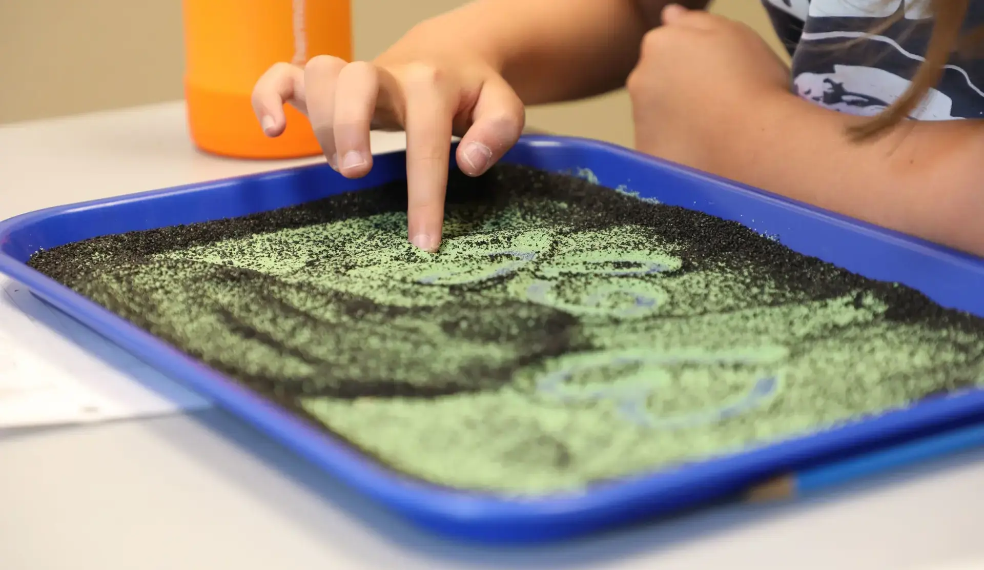 Child tracing shapes with a finger in a sensory sand tray.