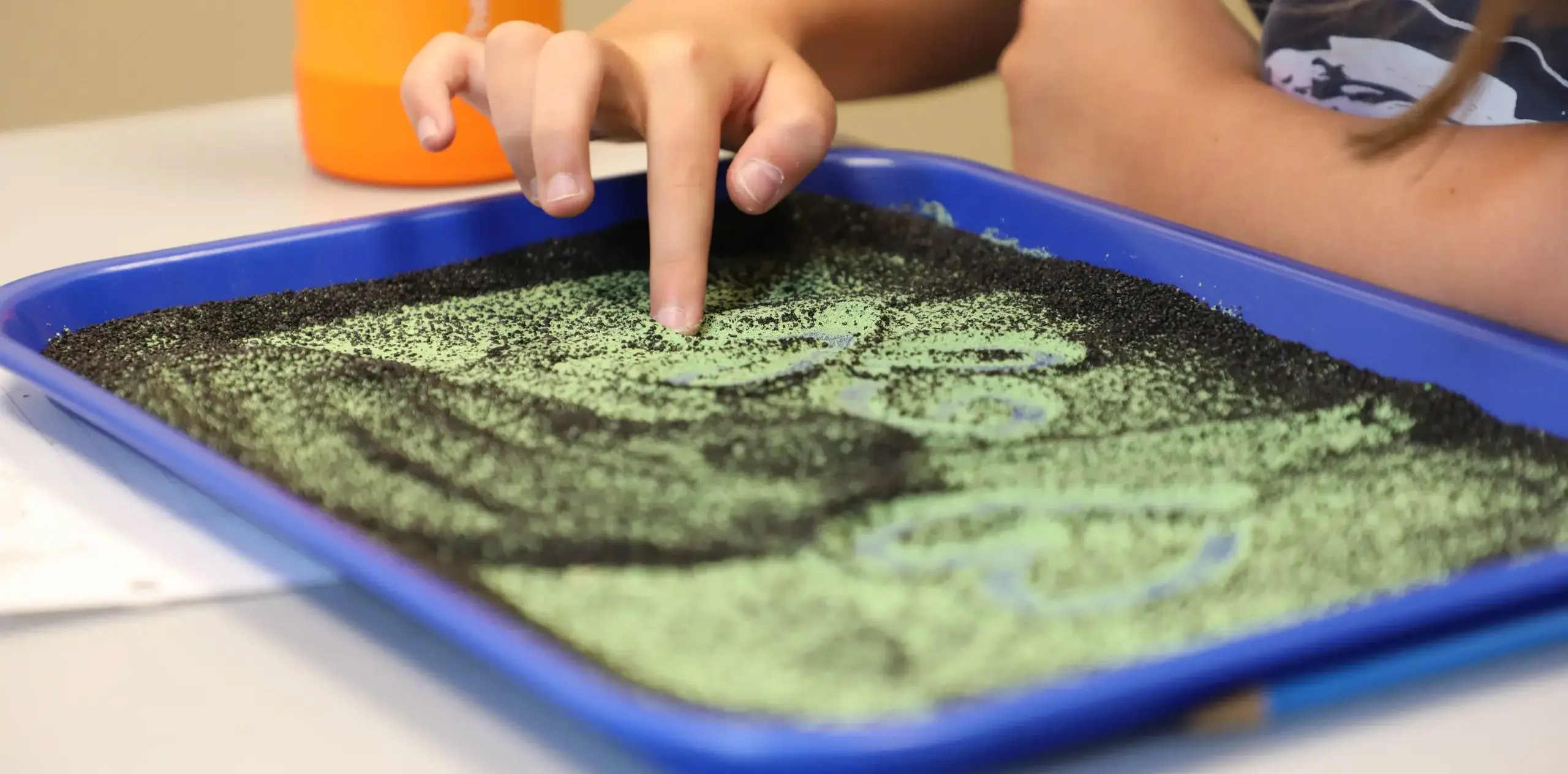 Child tracing shapes with a finger in a sensory sand tray.