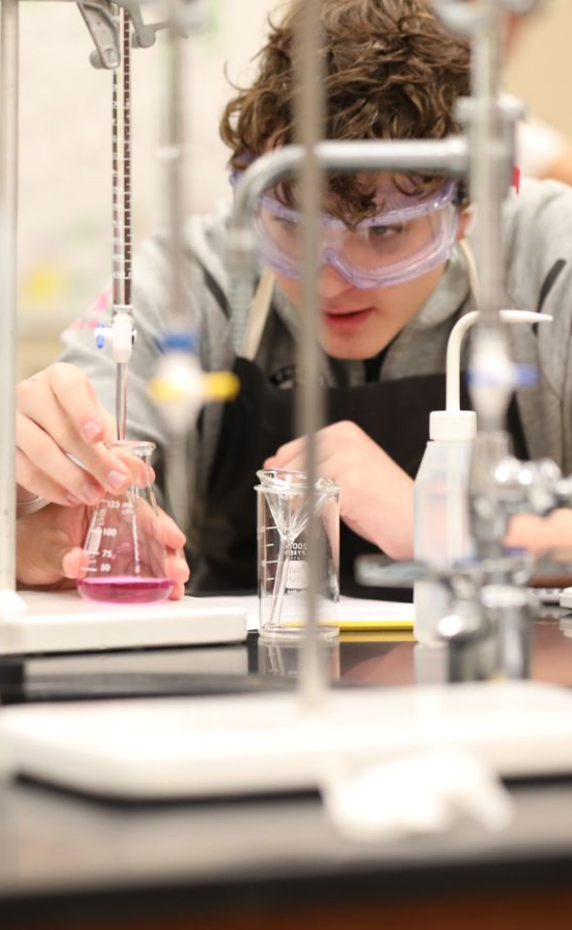 tudent wearing safety goggles conducts a chemistry experiment with laboratory glassware.