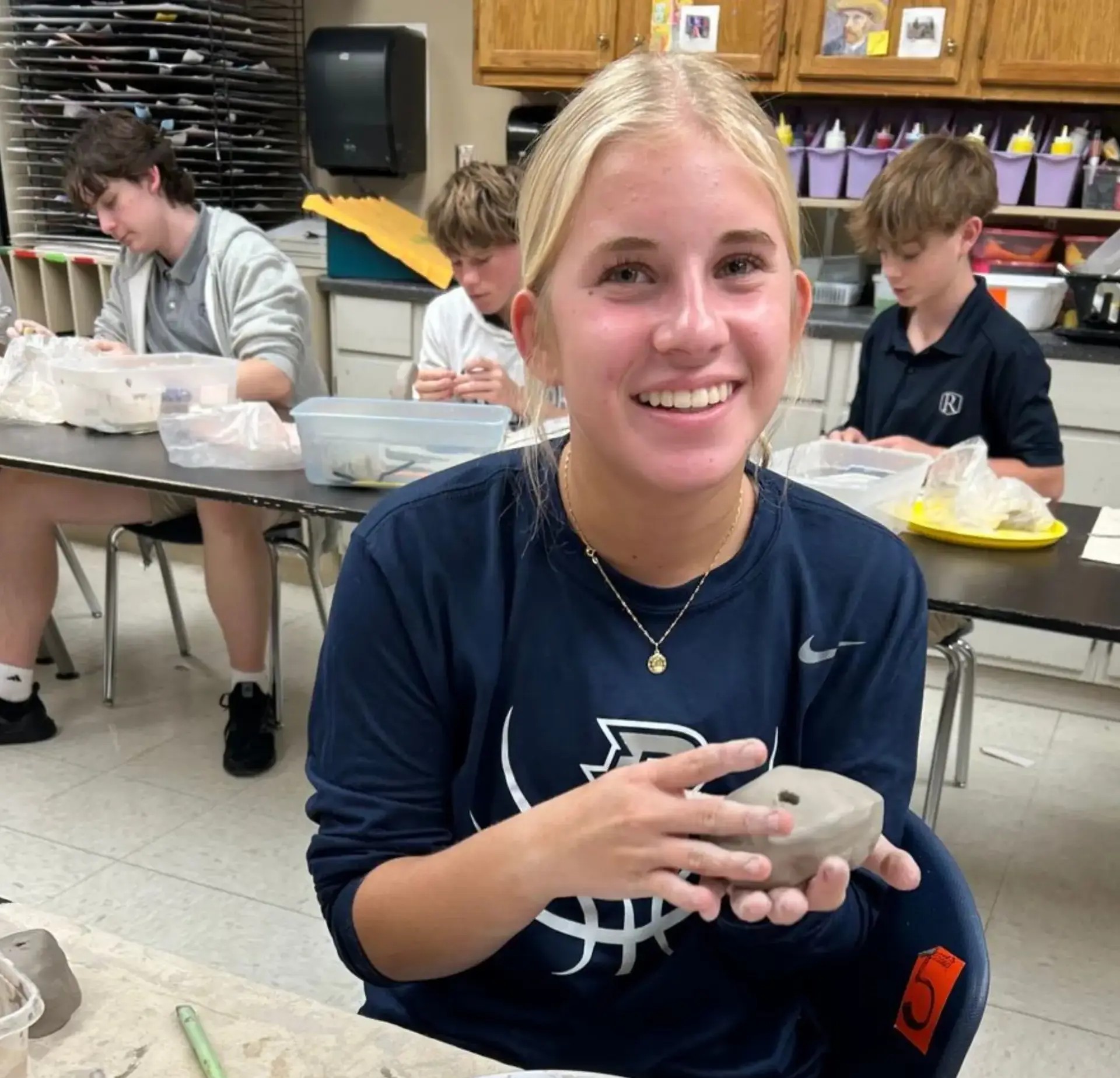 Smiling student holds a handmade clay project during an art class while classmates work in the background.