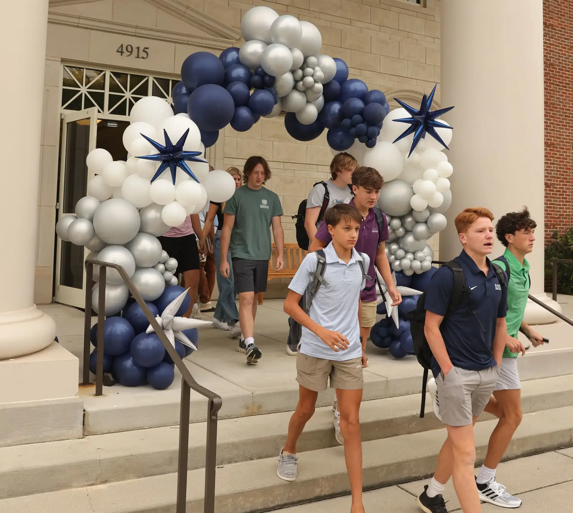 Students walk through a blue, white, and silver balloon arch outside a school entrance.
